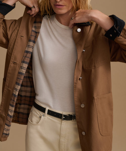 A model wearing a tan jacket over a cream t-shirt and beige trousers, with a plaid lining visible inside the jacket