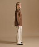 A woman stands in profile wearing a brown cropped jacket with a black collar, cream wide-leg trousers, and black shoes, photographed against a plain warm beige studio background.