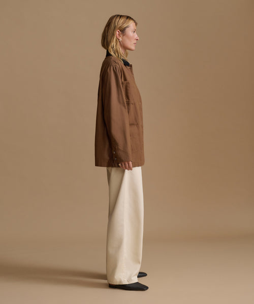 A woman stands in profile wearing a brown cropped jacket with a black collar, cream wide-leg trousers, and black shoes, photographed against a plain warm beige studio background.