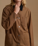 A close-up of a model wearing a brown utility shirt jacket with a black collar, silver buttons, and front pockets, set against a solid beige background.