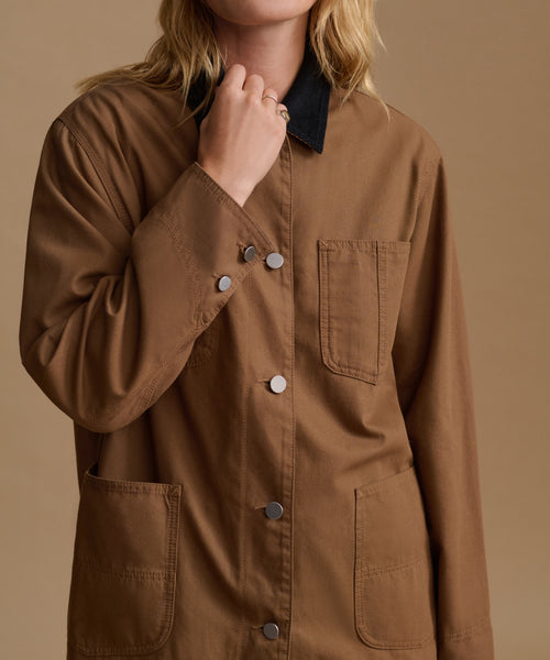 A close-up of a model wearing a brown utility shirt jacket with a black collar, silver buttons, and front pockets, set against a solid beige background.