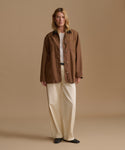 A woman stands against a plain beige studio background wearing a brown button-front jacket over a white top, cream wide-leg pants, and black shoes.