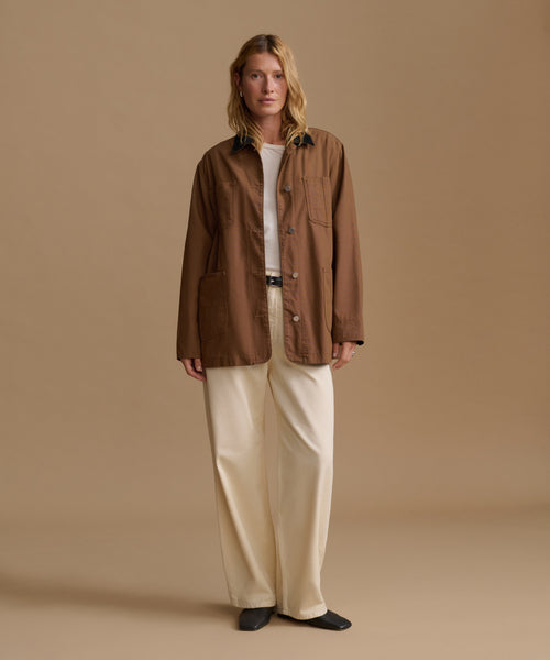 A woman stands against a plain beige studio background wearing a brown button-front jacket over a white top, cream wide-leg pants, and black shoes.