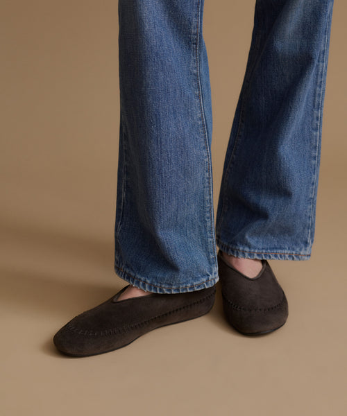 A close-up of a person's feet wearing dark brown suede moccasin-style slip-on shoes with blue jeans, set against a solid warm beige background.