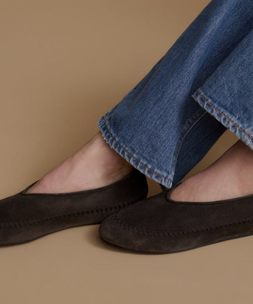 A close-up of a person wearing dark brown suede moccasin-style flats with blue jeans, set against a solid tan background.