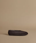 A single dark brown suede slip-on moccasin-style shoe with decorative stitching and a gray plush lining, shown in profile against a solid beige background.