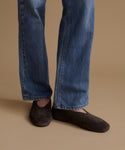 Close-up of a person's legs wearing blue jeans and dark brown suede slip-on shoes against a solid beige background.