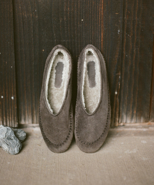 A pair of suede slip-on moccasin-style slippers with cream sherpa lining, displayed on a light concrete floor against a dark wooden plank background.