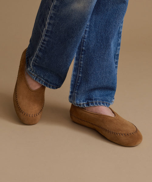 A person wearing brown suede moccasin-style slip-on shoes with contrast stitching, paired with blue jeans, against a solid beige studio background.