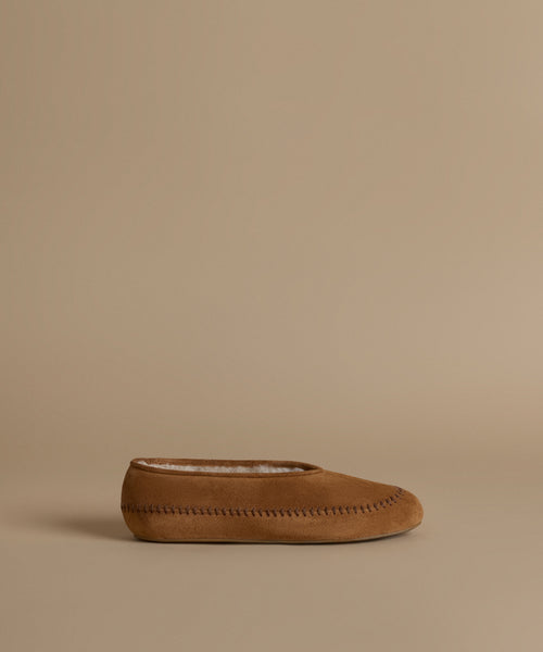 A single tan suede moccasin slipper with contrast stitching and a white lining, shown in profile against a solid beige background.
