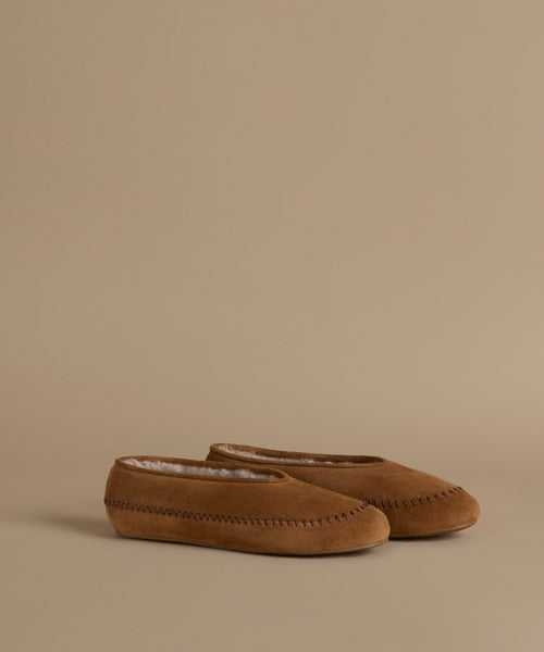 A pair of tan suede slip-on slippers with whipstitching and a soft white lining, displayed on a solid beige background.