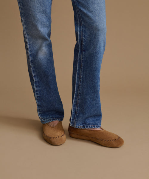 A person wearing blue jeans and tan suede moccasin-style slip-on shoes, shown from the knees down against a plain warm beige studio background.