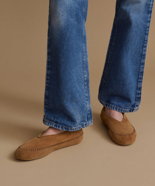 A person wearing blue jeans and tan suede slip-on moccasin-style shoes against a plain beige studio background.