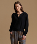 A woman modeling a black long-sleeve button-front cardigan with a relaxed fit, paired with brown and beige vertically striped pants, against a solid warm tan studio background.
