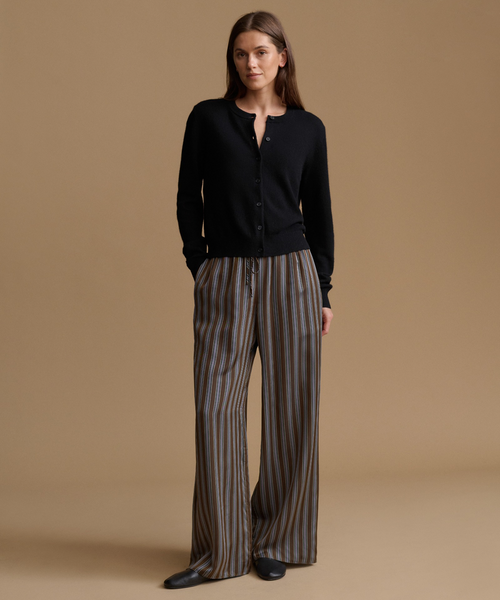 A woman stands against a warm beige studio background wearing a black long-sleeve button-front cardigan and wide-leg striped trousers in brown, gray, and beige tones, paired with black flats.