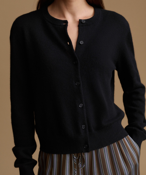 A close-up of a person wearing a black cardigan with a buttoned front and ribbed collar, cuffs, and hem, paired with a striped skirt against a warm tan background.