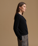 A woman in side profile wearing a black button-front cardigan and striped trousers, standing against a solid warm tan studio background.