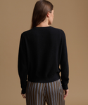 A woman shown from the back wearing a black long-sleeve knit sweater and brown, beige, and gray vertically striped trousers, set against a solid warm tan studio background.