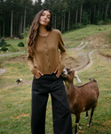 A woman stands outdoors in a grassy field with trees in the background, wearing a brown button-front cardigan and black wide-leg pants, with a goat beside her.