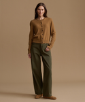 A woman stands against a warm tan studio background wearing a camel ribbed knit cardigan, olive green wide-leg trousers, and tan suede shoes.