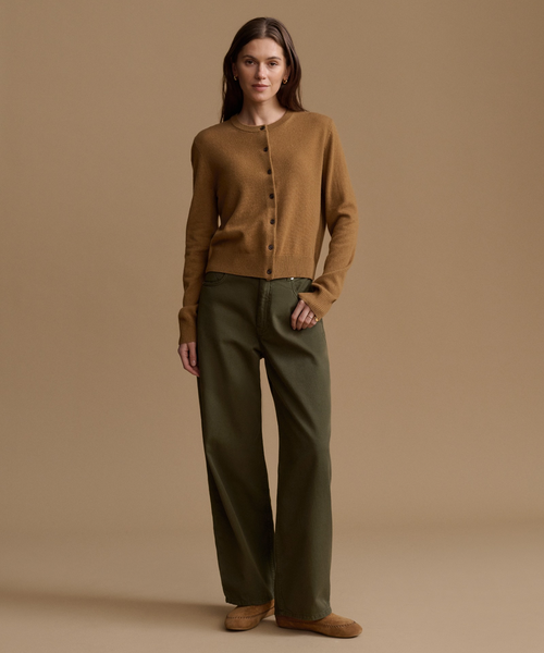 A woman stands against a warm tan studio background wearing a camel ribbed knit cardigan, olive green wide-leg trousers, and tan suede shoes.