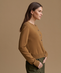 A woman in side profile wearing a camel brown button-up cardigan sweater and olive green pants, set against a solid warm tan studio background.