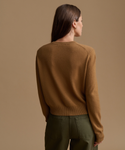 A woman shown from the back wearing a camel brown knit sweater and olive green pants, set against a solid warm beige studio background.
