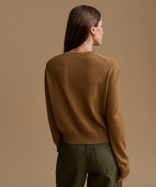 A woman shown from the back wearing a camel brown knit sweater and olive green pants, set against a solid warm beige studio background.