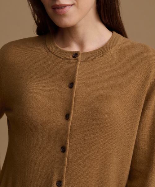 Close-up of a person wearing a warm camel-colored knit cardigan sweater with dark buttons, against a soft beige background.