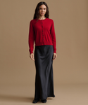 A woman stands against a plain warm beige studio background wearing a bright red long-sleeve button-front cardigan and a long black satin-like skirt.