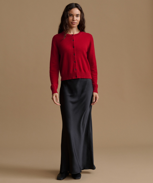 A woman stands against a plain warm beige studio background wearing a bright red long-sleeve button-front cardigan and a long black satin-like skirt.