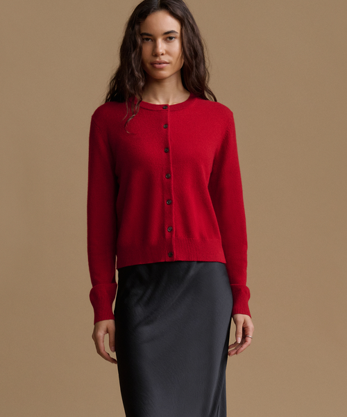 A woman modeling a bright red long-sleeve knit cardigan with dark buttons, paired with a black skirt, against a solid beige studio background.