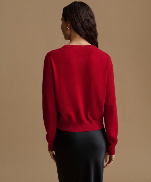 A woman shown from the back wearing a bright red knit sweater and a black skirt, standing against a plain beige studio background.