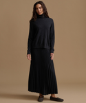 A woman stands against a plain warm tan studio background wearing a dark charcoal turtleneck sweater and a long black pleated skirt, with dark slip-on shoes.