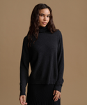A woman modeling a dark charcoal gray turtleneck sweater against a solid tan studio background.