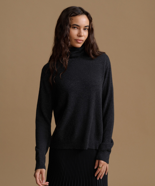 A woman modeling a dark charcoal gray turtleneck sweater against a solid tan studio background.
