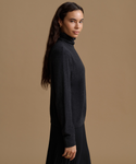 A woman wearing a dark charcoal gray ribbed turtleneck sweater and a black pleated skirt, shown in profile against a solid warm beige studio background.