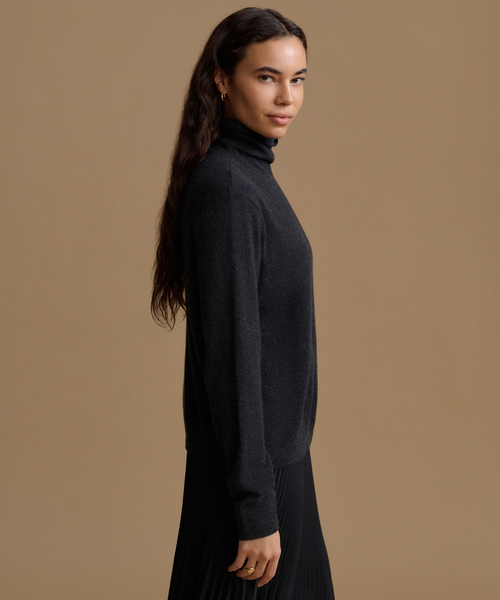 A woman wearing a dark charcoal gray ribbed turtleneck sweater and a black pleated skirt, shown in profile against a solid warm beige studio background.