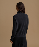 A woman shown from the back wearing a dark charcoal gray turtleneck sweater and a black pleated skirt, set against a solid warm beige studio background.