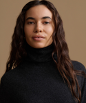 A close-up portrait of a woman with long, wavy dark brown hair wearing a dark charcoal gray turtleneck sweater against a solid warm beige background.