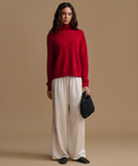 A woman stands against a plain warm beige studio background wearing a bright red mock-neck knit sweater, cream wide-leg pants, black shoes, and holding a small black woven handbag.