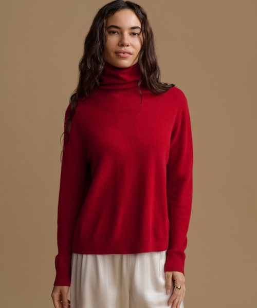 A woman wearing a vibrant red knit turtleneck sweater and cream-colored trousers, standing against a solid warm brown studio background.