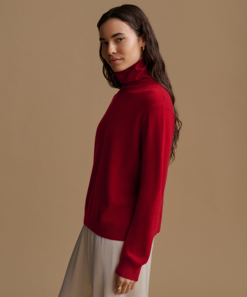 A woman wearing a vibrant red turtleneck sweater and light beige trousers, posed in profile against a solid warm beige studio background.