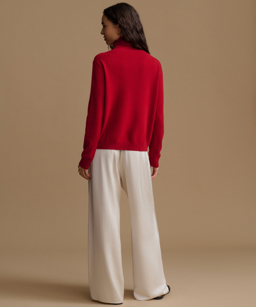 A woman shown from the back wearing a bright red mock-neck knit sweater and wide-leg cream trousers, standing against a solid beige studio background.