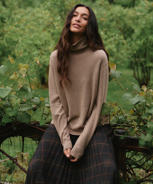 A woman wearing a beige cowl-neck knit sweater and a dark plaid pleated skirt, sitting outdoors on a wooden fence with green garden foliage in the background.