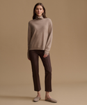 A woman stands against a warm beige studio background wearing a taupe turtleneck sweater, dark brown straight-leg trousers, and taupe slip-on shoes.