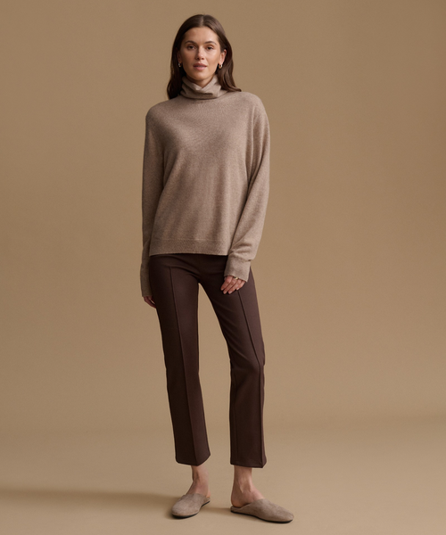 A woman stands against a warm beige studio background wearing a taupe turtleneck sweater, dark brown straight-leg trousers, and taupe slip-on shoes.