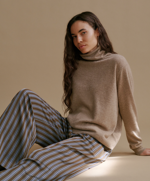 A woman wearing a taupe brown turtleneck sweater and striped trousers, sitting against a solid beige background.