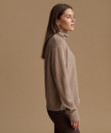 A woman in side profile wearing a light taupe knit turtleneck sweater and dark brown pants, set against a solid warm tan studio background.