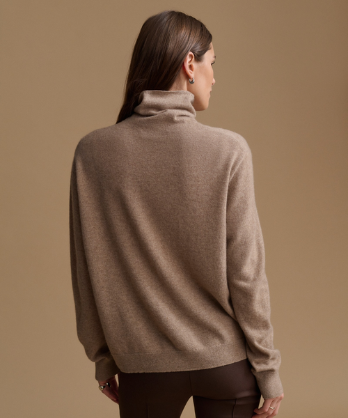 A woman shown from the back wearing a taupe brown turtleneck sweater and dark brown pants, set against a solid warm beige studio background.